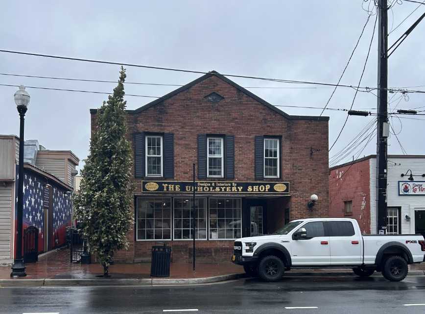 Brick storefront with a two-story facade and a sign reading The Upholstery Shop; a white pickup truck is parked on a wet street in front on a cloudy day.