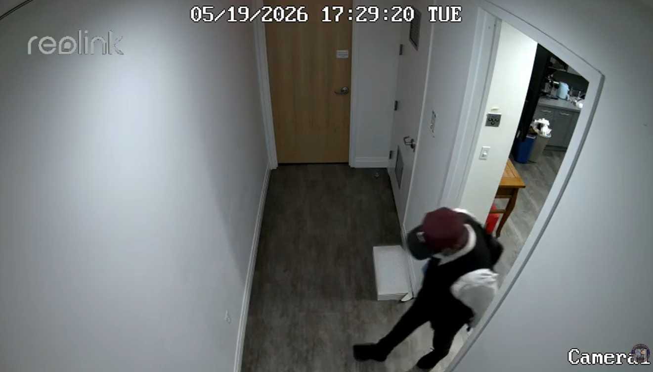 Security camera footage showing a person in a burgundy hat and dark clothing walking down a narrow hallway toward a wooden door at the end of the hall.