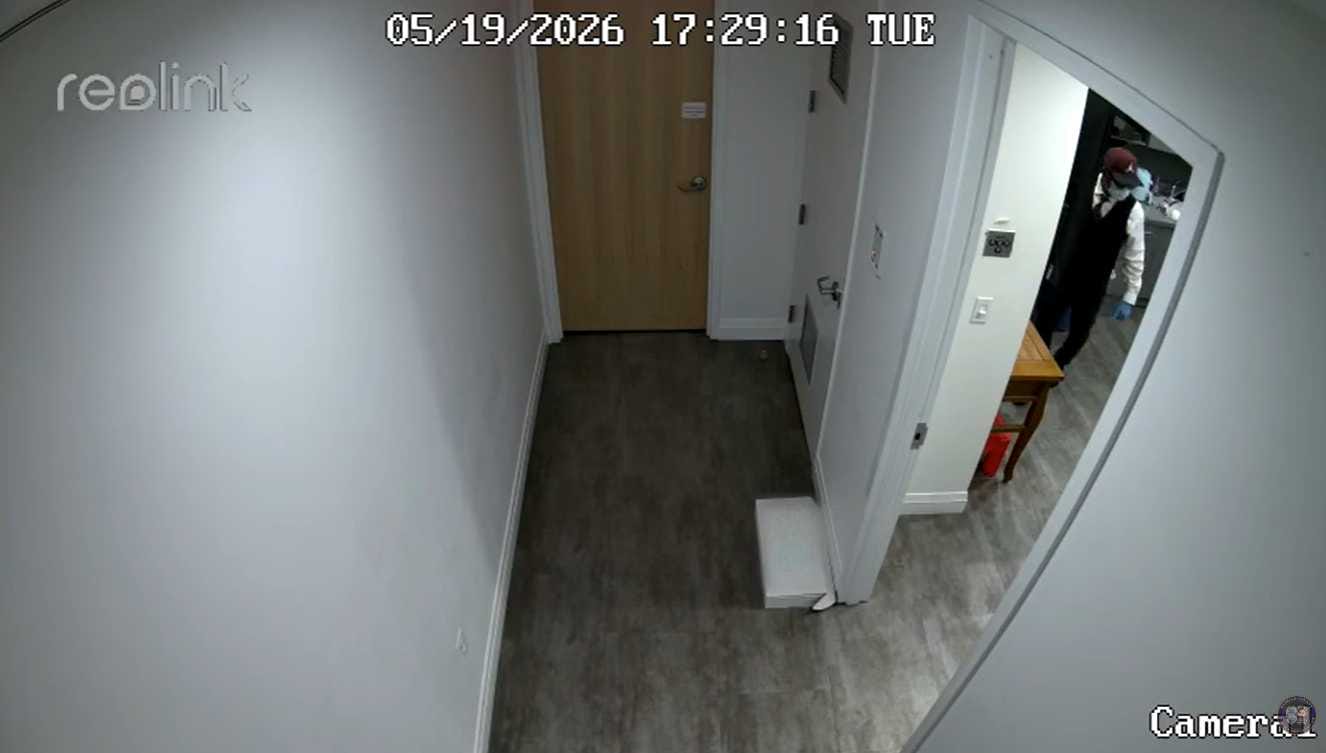 Fisheye surveillance view of a narrow hallway with a closed wooden door at the end and a man in a vest and red cap standing near a small wooden table in the doorway on the right.