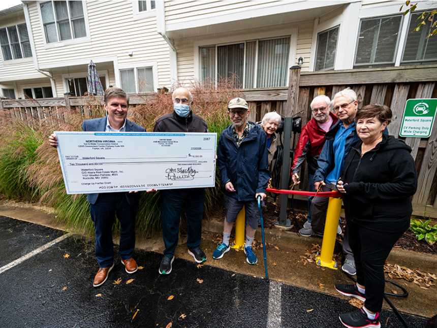 Hunter Mill District Supervisor Walter Alcorn and Waterford Square Condominium Association residents celebrate the installation of an electric vehicle charger (via Hunter Mill District Office) | FFXnow Eight people stand outside townhouses, presenting a large ceremonial check during a donation event.
