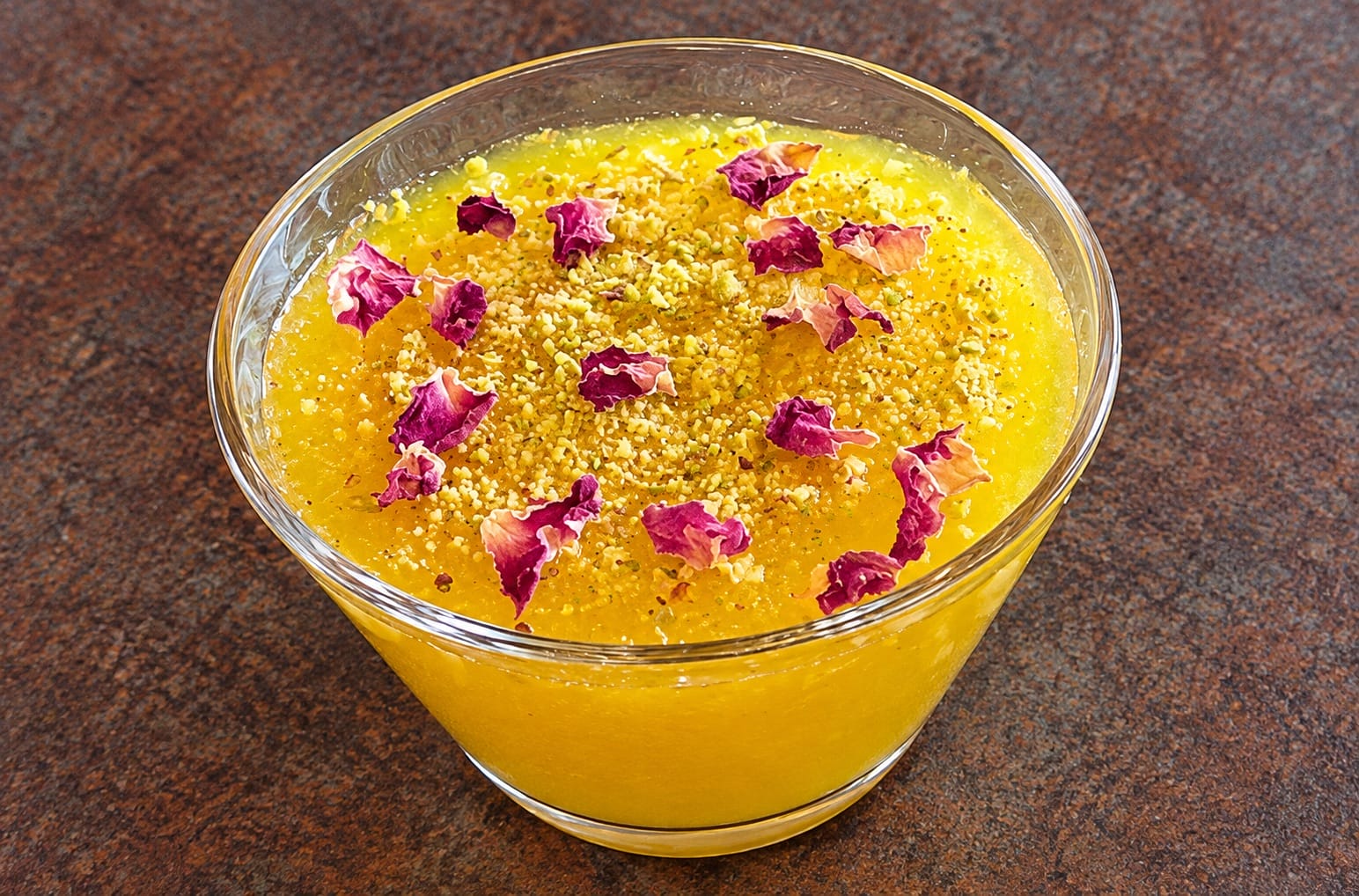 Bright yellow mango dessert in a glass bowl topped with crushed pistachios and pink rose petals, on a dark countertop, close-up.