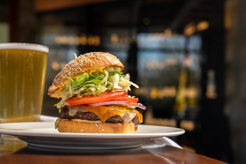 Burger with sesame bun, lettuce, tomato, onions and cheese on a plate, with a glass of beer beside it.