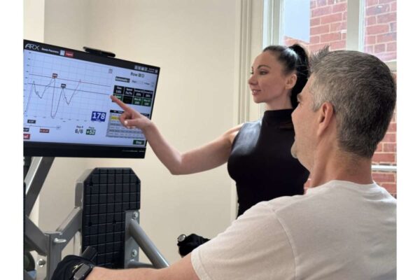 Fitness trainer explains workout data on a large screen to a man seated on gym equipment.