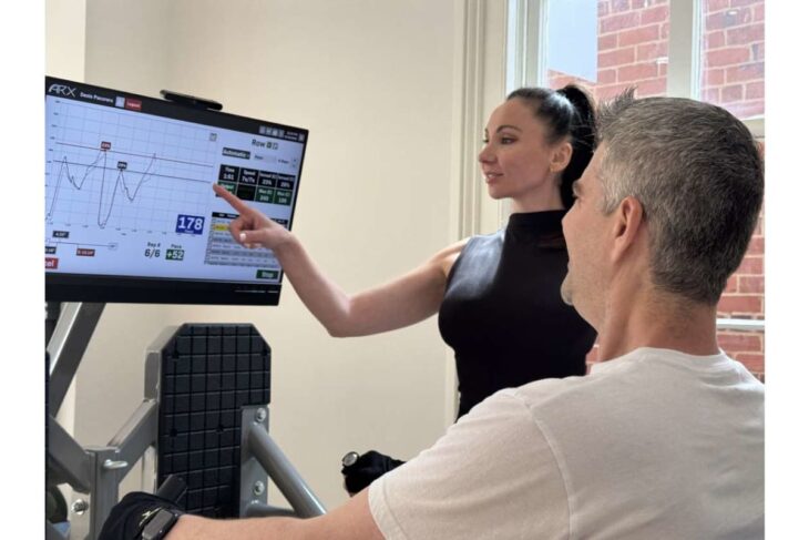 Fitness trainer explains workout data on a large screen to a man seated on gym equipment.