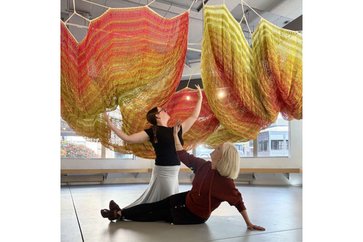 Two people in a studio interact with a large, colorful fabric sculpture hanging overhead; a standing woman reaches up while a seated woman supports the fabric.