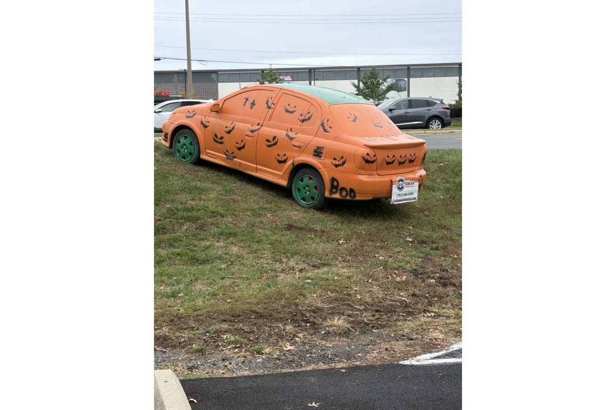 Orange car painted with black jack-o'-lantern faces and green wheels, parked on grass in a lot (Halloween-themed).