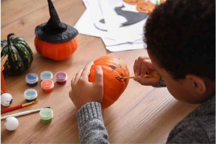 Child decorating a small orange pumpkin with a pencil/marker while paint pots and brushes sit on a wooden table.
