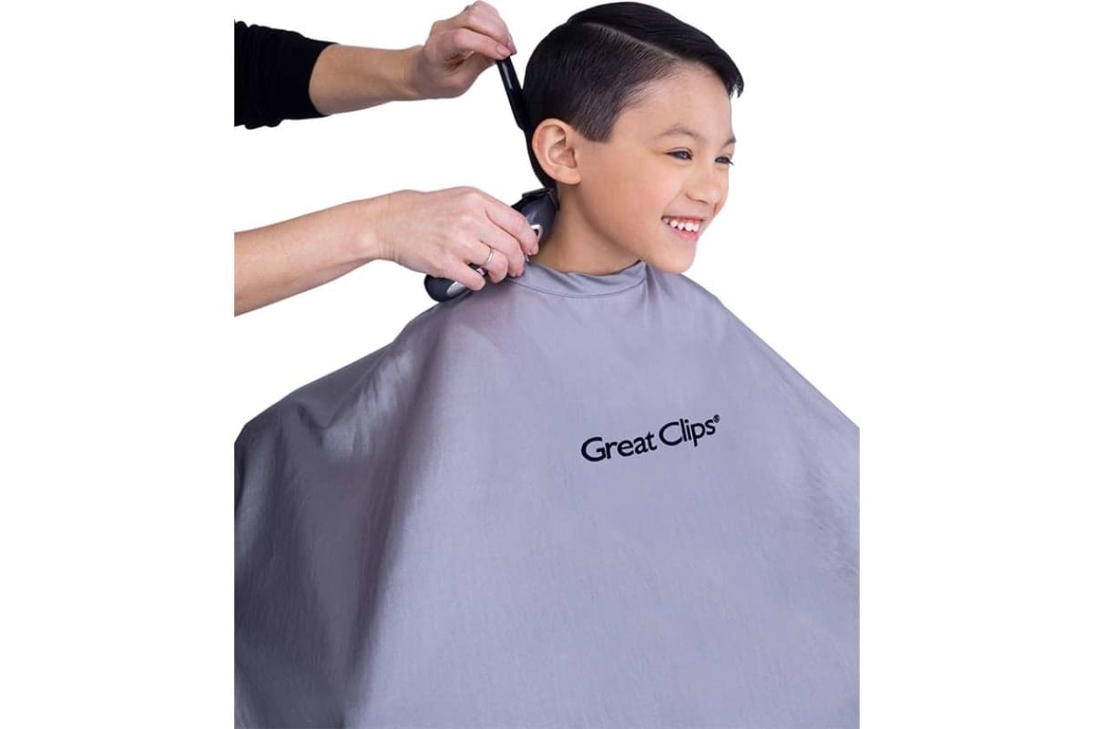 Child smiling as a barber trims hair on the neck; wearing a gray Great Clips cape.