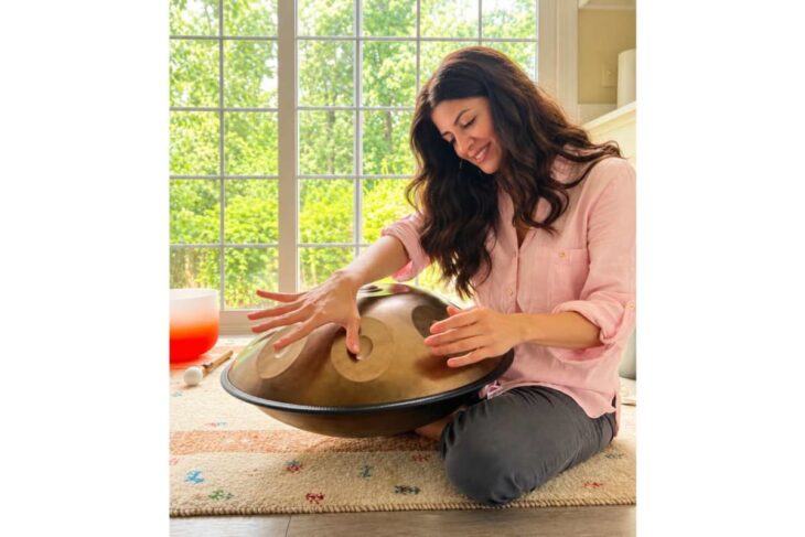 Woman in a pink shirt sits on a rug, playing a handpan near a bright window.