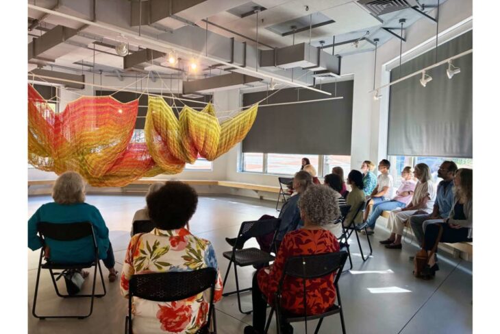 Audience seated in a bright studio watching a large orange‑yellow fabric sculpture suspended from the ceiling.